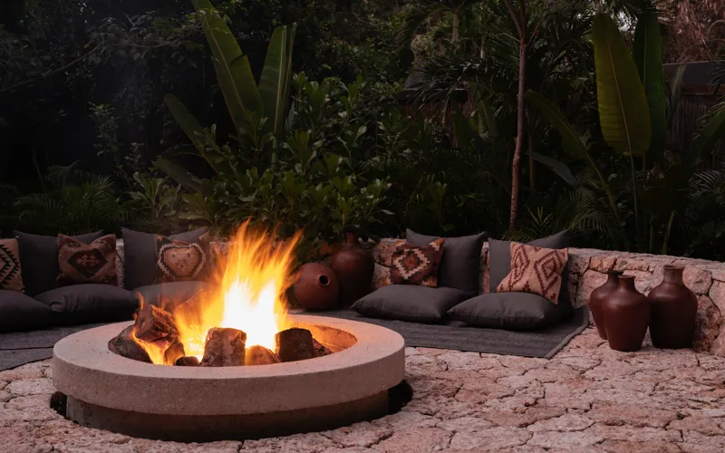 A beautifully designed outdoor lounge area at La Reserva Tulum, featuring dark walls, a tree growing through the floor, stylish dark furniture, a round coffee table, and woven pendant lights, all set against a backdrop of lush tropical foliage wit...