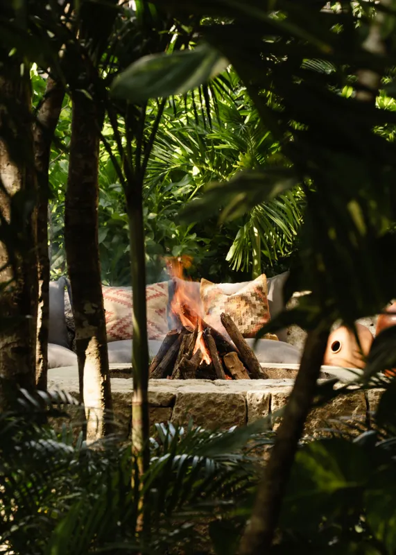 A beautifully designed outdoor lounge area at La Reserva Tulum, featuring dark walls, a tree growing through the floor, stylish dark furniture, a round coffee table, and woven pendant lights, all set against a backdrop of lush tropical foliage wit...