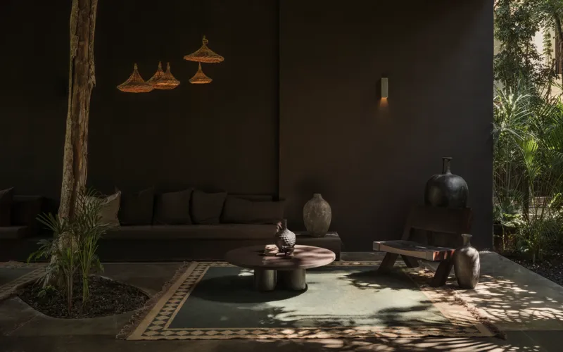 A beautifully designed outdoor lounge area at La Reserva Tulum, featuring dark walls, a tree growing through the floor, stylish dark furniture, a round coffee table, and woven pendant lights, all set against a backdrop of lush tropical foliage wit...