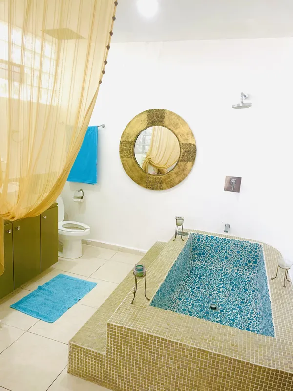 A bright and inviting living room in Casa Dragonfly featuring a white sectional sofa with colorful Moroccan-inspired pillows, a wooden coffee table with a decorative lantern, a round patterned floor cushion, and a large ornate window, showcasing t...