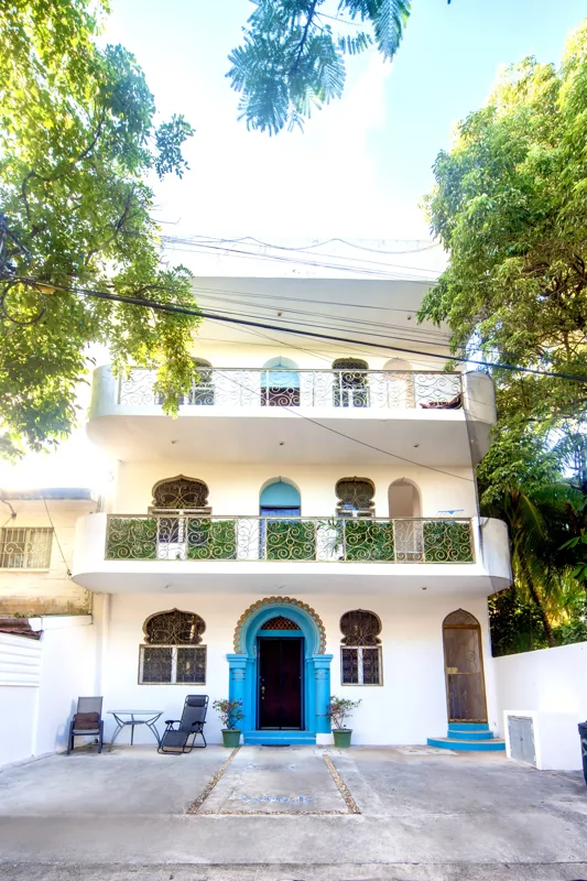 A bright and inviting living room in Casa Dragonfly featuring a white sectional sofa with colorful Moroccan-inspired pillows, a wooden coffee table with a decorative lantern, a round patterned floor cushion, and a large ornate window, showcasing t...