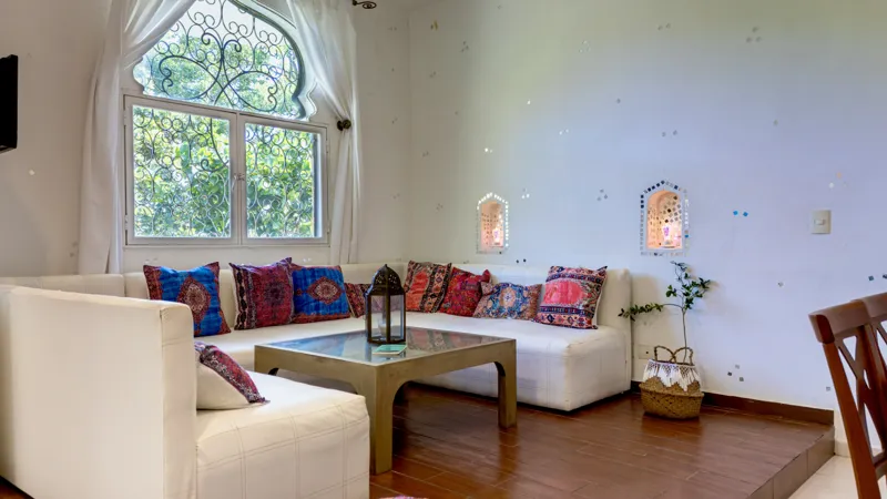 A bright and inviting living room in Casa Dragonfly featuring a white sectional sofa with colorful Moroccan-inspired pillows, a wooden coffee table with a decorative lantern, a round patterned floor cushion, and a large ornate window, showcasing t...