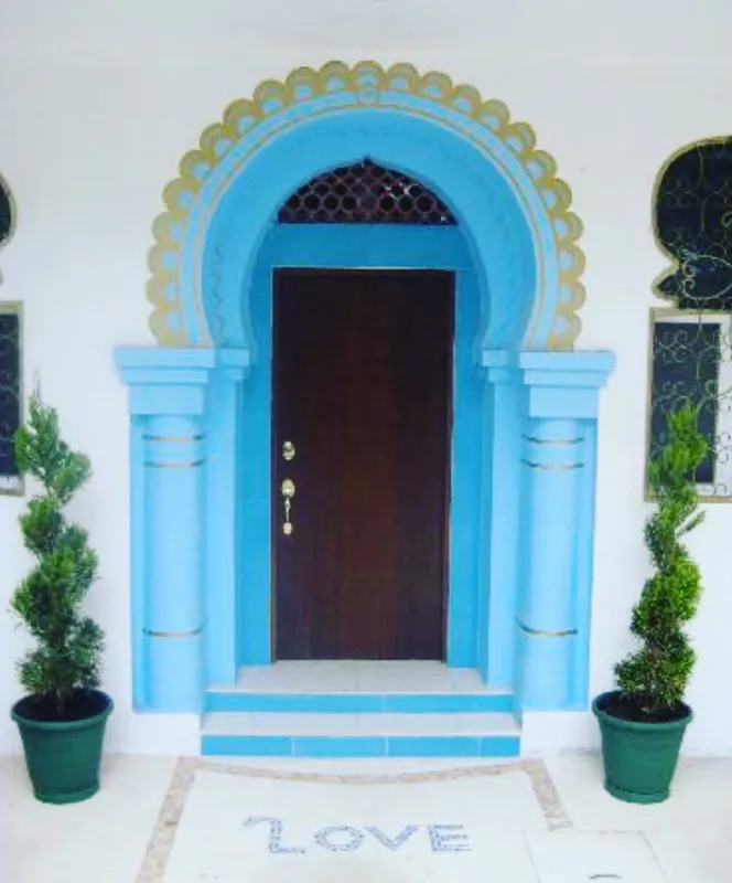 A bright and inviting living room in Casa Dragonfly featuring a white sectional sofa with colorful Moroccan-inspired pillows, a wooden coffee table with a decorative lantern, a round patterned floor cushion, and a large ornate window, showcasing t...