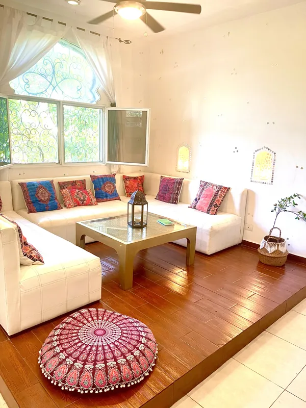 A bright and inviting living room in Casa Dragonfly featuring a white sectional sofa with colorful Moroccan-inspired pillows, a wooden coffee table with a decorative lantern, a round patterned floor cushion, and a large ornate window, showcasing t...