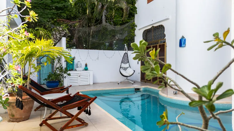A bright and inviting living room in Casa Dragonfly featuring a white sectional sofa with colorful Moroccan-inspired pillows, a wooden coffee table with a decorative lantern, a round patterned floor cushion, and a large ornate window, showcasing t...