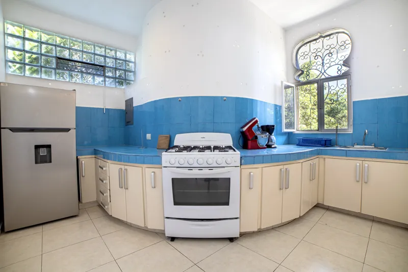A bright and inviting living room in Casa Dragonfly featuring a white sectional sofa with colorful Moroccan-inspired pillows, a wooden coffee table with a decorative lantern, a round patterned floor cushion, and a large ornate window, showcasing t...