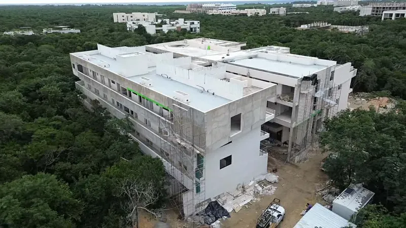 Aerial view of the Zamajal development in Tulum, showcasing multiple buildings in various stages of construction, with a construction vehicle on the ground and lush green jungle surrounding the site.