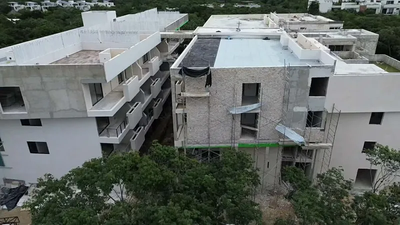 Aerial view of the Zamajal development in Tulum, showcasing multiple buildings in various stages of construction, with a construction vehicle on the ground and lush green jungle surrounding the site.