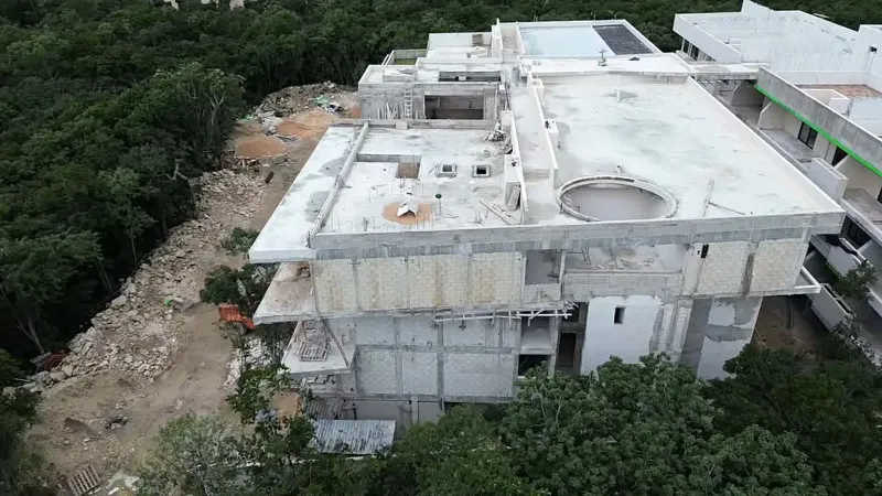 Aerial view of the Zamajal development in Tulum, showcasing multiple buildings in various stages of construction, with a construction vehicle on the ground and lush green jungle surrounding the site.
