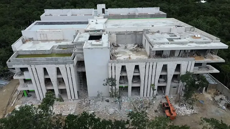 Aerial view of the Zamajal development in Tulum, showcasing multiple buildings in various stages of construction, with a construction vehicle on the ground and lush green jungle surrounding the site.