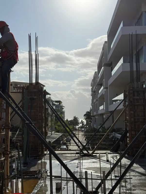 An active construction site for Costera Mamitas under a bright blue sky, featuring multiple workers in safety gear, rebar extending upwards, and wooden concrete forms, indicating significant structural progress.