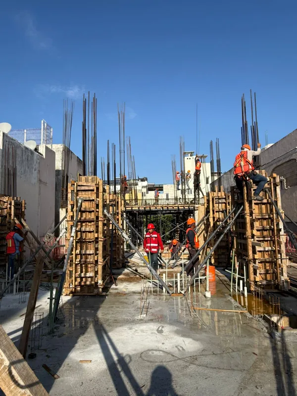 An active construction site for Costera Mamitas under a bright blue sky, featuring multiple workers in safety gear, rebar extending upwards, and wooden concrete forms, indicating significant structural progress.