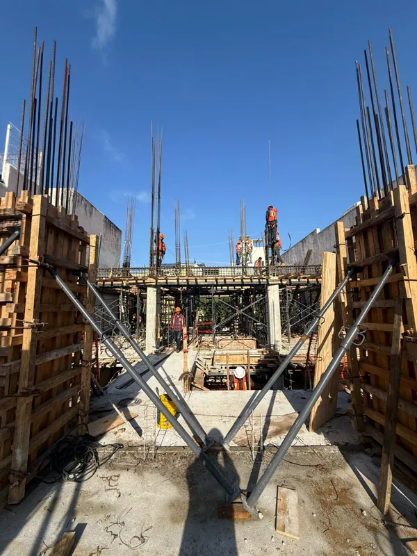 An active construction site for Costera Mamitas under a bright blue sky, featuring multiple workers in safety gear, rebar extending upwards, and wooden concrete forms, indicating significant structural progress.