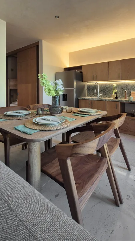 A beautifully furnished dining area with a wooden table set for four, surrounded by modern wooden chairs, leading into a contemporary kitchen with dark wood cabinets, light granite countertops, and stainless steel appliances, all under warm accent...