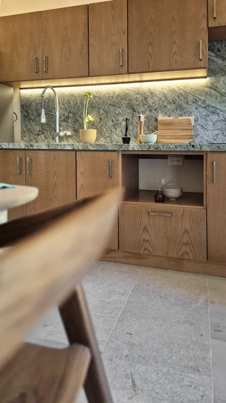 A beautifully furnished dining area with a wooden table set for four, surrounded by modern wooden chairs, leading into a contemporary kitchen with dark wood cabinets, light granite countertops, and stainless steel appliances, all under warm accent...