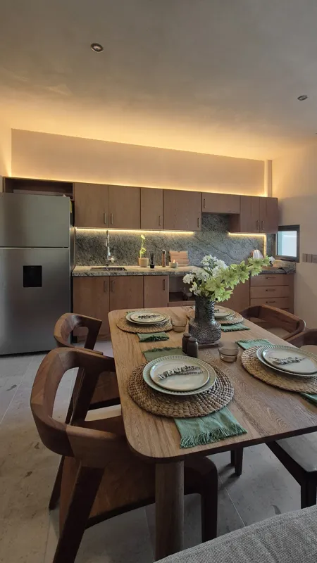 A beautifully furnished dining area with a wooden table set for four, surrounded by modern wooden chairs, leading into a contemporary kitchen with dark wood cabinets, light granite countertops, and stainless steel appliances, all under warm accent...