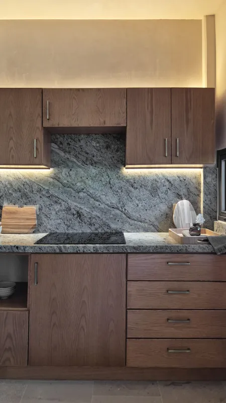 A beautifully furnished dining area with a wooden table set for four, surrounded by modern wooden chairs, leading into a contemporary kitchen with dark wood cabinets, light granite countertops, and stainless steel appliances, all under warm accent...