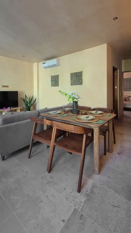 A beautifully furnished dining area with a wooden table set for four, surrounded by modern wooden chairs, leading into a contemporary kitchen with dark wood cabinets, light granite countertops, and stainless steel appliances, all under warm accent...