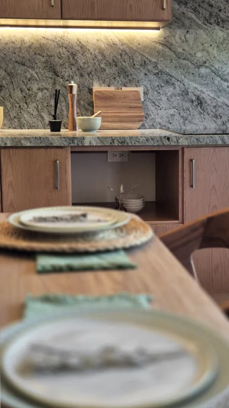 A beautifully furnished dining area with a wooden table set for four, surrounded by modern wooden chairs, leading into a contemporary kitchen with dark wood cabinets, light granite countertops, and stainless steel appliances, all under warm accent...