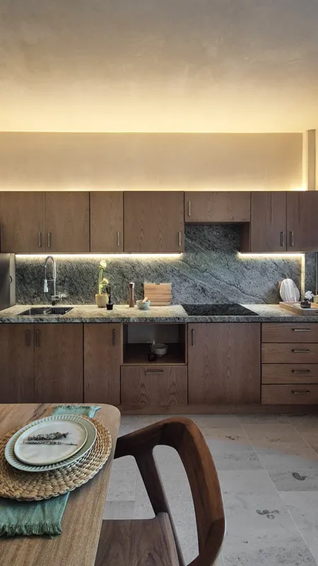 A beautifully furnished dining area with a wooden table set for four, surrounded by modern wooden chairs, leading into a contemporary kitchen with dark wood cabinets, light granite countertops, and stainless steel appliances, all under warm accent...