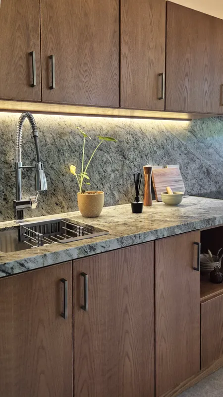 A beautifully furnished dining area with a wooden table set for four, surrounded by modern wooden chairs, leading into a contemporary kitchen with dark wood cabinets, light granite countertops, and stainless steel appliances, all under warm accent...