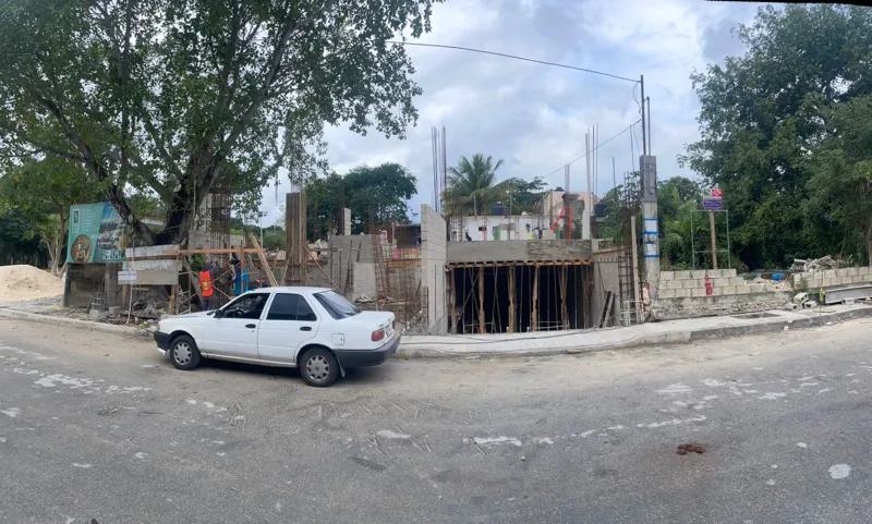 Panoramic view of the Polo 5ta construction site in Playa del Carmen, showing the initial stages of the building's concrete structure with columns and slabs, surrounded by lush trees and a street in the foreground. A white sedan is parked on the l...