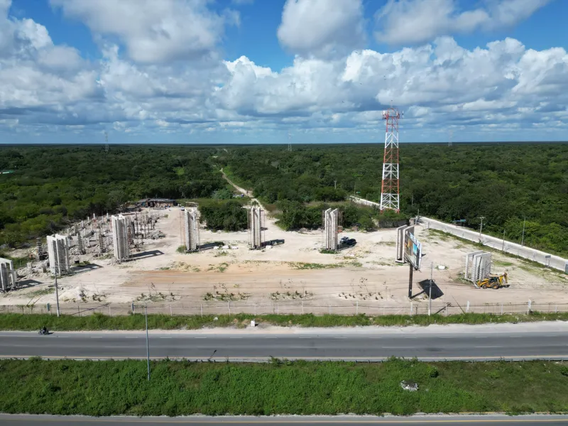Aerial view of the Anthar development construction site in Playa del Carmen, showing numerous concrete pillars and walls erected, alongside ongoing foundation work and construction vehicles, with a highway and lush green landscape in the backgroun...