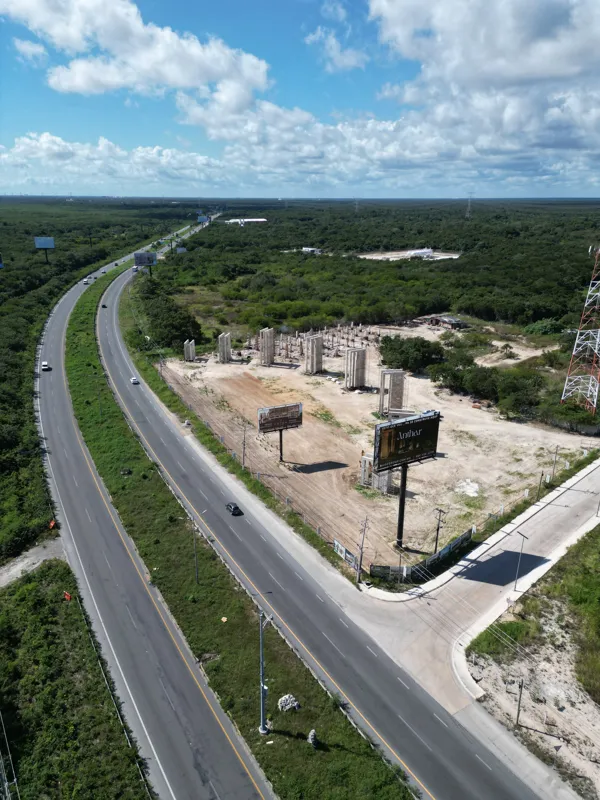 Aerial view of the Anthar development construction site in Playa del Carmen, showing numerous concrete pillars and walls erected, alongside ongoing foundation work and construction vehicles, with a highway and lush green landscape in the backgroun...