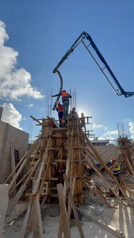 Construction workers in safety gear are actively pouring concrete for the Spirit 1990 development, using a large concrete pump with its boom extended over the building structure, which is supported by intricate wooden formwork and visible rebar un...