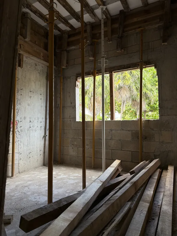 An aerial view from a partially constructed building at Soulam, showing rebar extending upwards, concrete block walls, and wooden formwork. In the background, a traditional thatched-roof building is visible amidst lush tropical greenery under a pa...