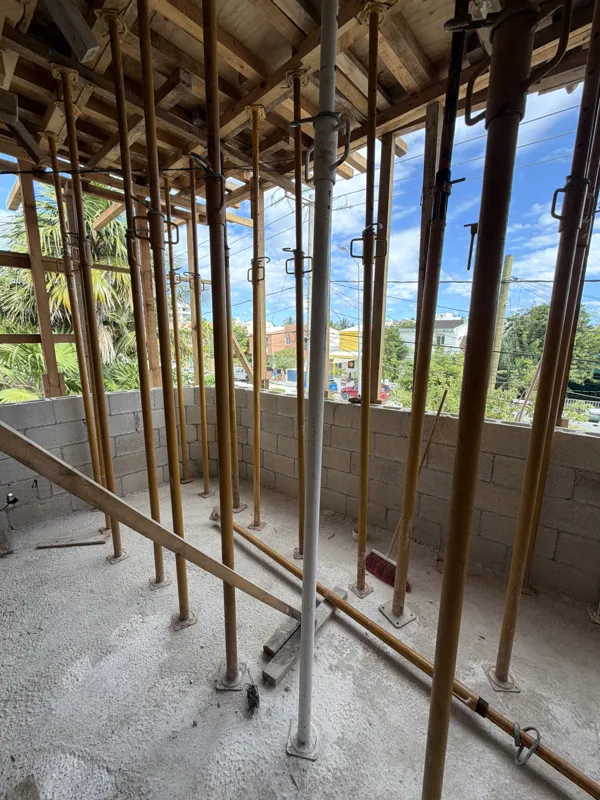 An aerial view from a partially constructed building at Soulam, showing rebar extending upwards, concrete block walls, and wooden formwork. In the background, a traditional thatched-roof building is visible amidst lush tropical greenery under a pa...