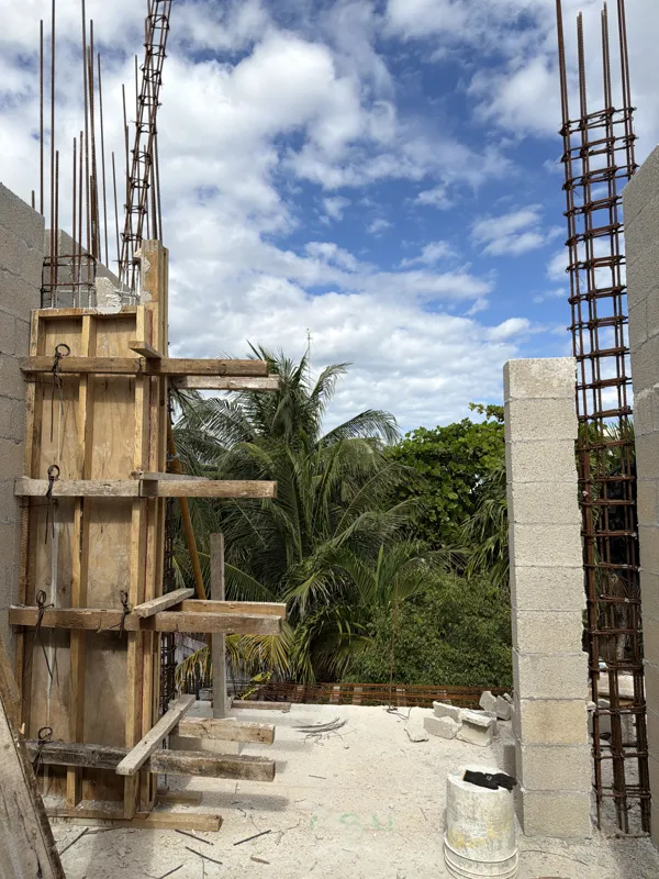 An aerial view from a partially constructed building at Soulam, showing rebar extending upwards, concrete block walls, and wooden formwork. In the background, a traditional thatched-roof building is visible amidst lush tropical greenery under a pa...