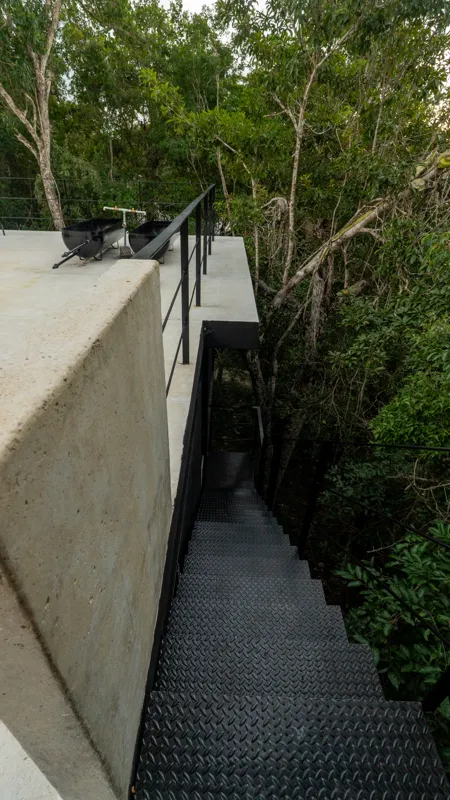 High-angle view of a modern living and kitchen area in a Tree60 2-bedroom Essence Jungle Villa, featuring large windows overlooking lush jungle, a light-colored sofa, dark kitchen island with stools, and concrete-style flooring.