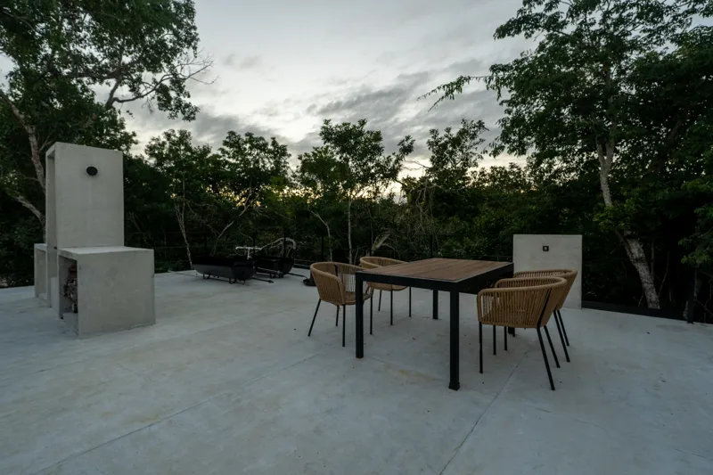 High-angle view of a modern living and kitchen area in a Tree60 2-bedroom Essence Jungle Villa, featuring large windows overlooking lush jungle, a light-colored sofa, dark kitchen island with stools, and concrete-style flooring.