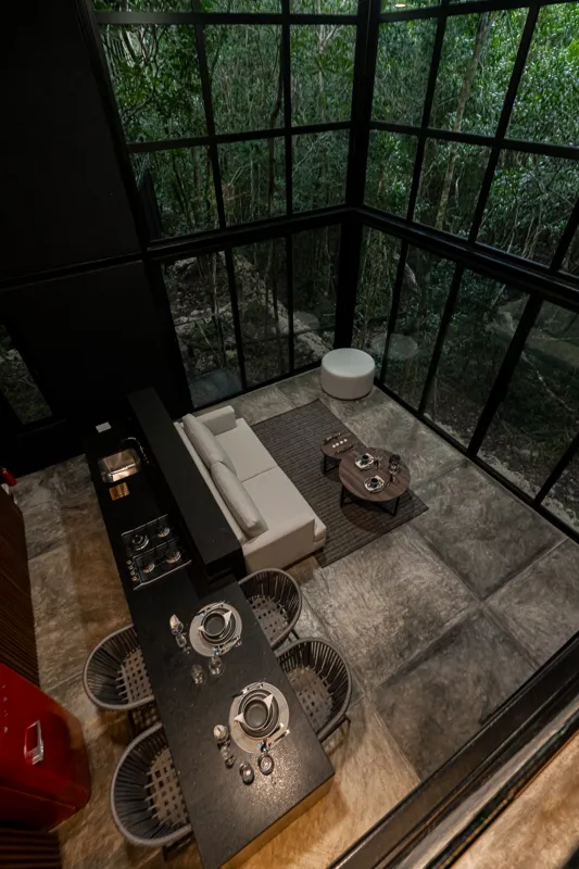 High-angle view of a modern living and kitchen area in a Tree60 2-bedroom Essence Jungle Villa, featuring large windows overlooking lush jungle, a light-colored sofa, dark kitchen island with stools, and concrete-style flooring.