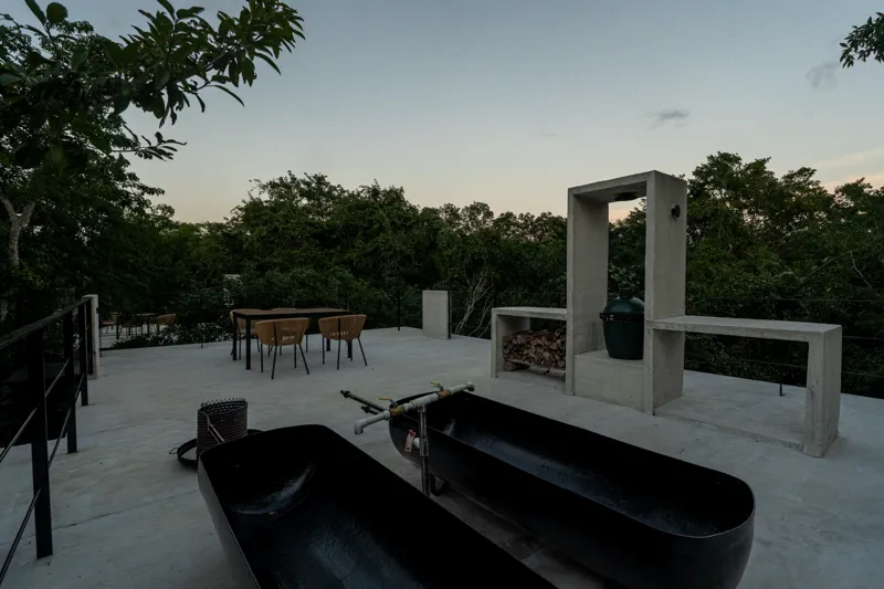 High-angle view of a modern living and kitchen area in a Tree60 2-bedroom Essence Jungle Villa, featuring large windows overlooking lush jungle, a light-colored sofa, dark kitchen island with stools, and concrete-style flooring.