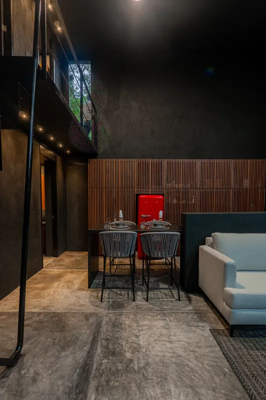 High-angle view of a modern living and kitchen area in a Tree60 2-bedroom Essence Jungle Villa, featuring large windows overlooking lush jungle, a light-colored sofa, dark kitchen island with stools, and concrete-style flooring.