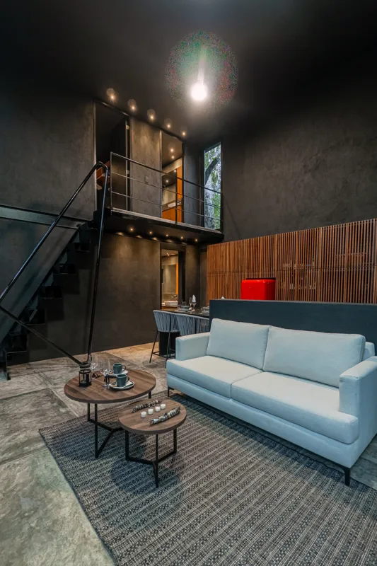 High-angle view of a modern living and kitchen area in a Tree60 2-bedroom Essence Jungle Villa, featuring large windows overlooking lush jungle, a light-colored sofa, dark kitchen island with stools, and concrete-style flooring.