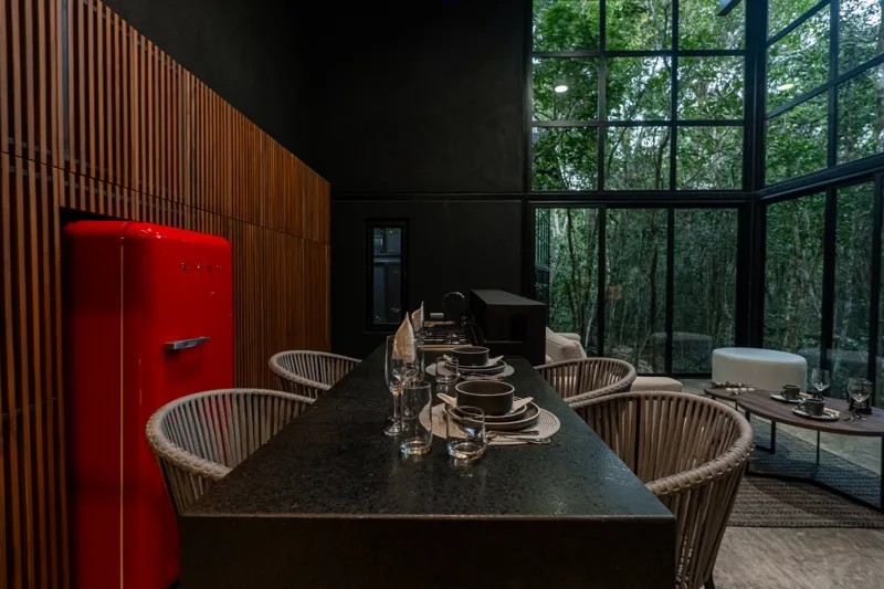 High-angle view of a modern living and kitchen area in a Tree60 2-bedroom Essence Jungle Villa, featuring large windows overlooking lush jungle, a light-colored sofa, dark kitchen island with stools, and concrete-style flooring.