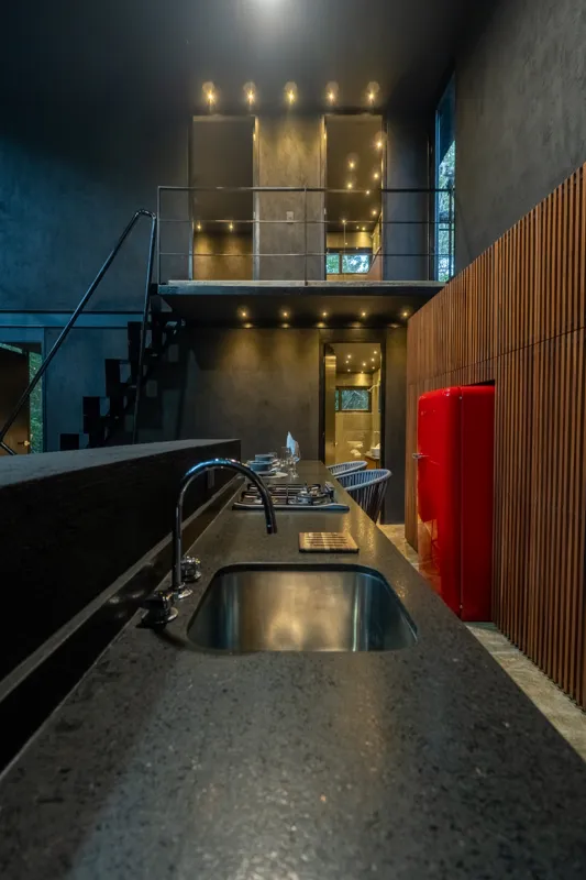High-angle view of a modern living and kitchen area in a Tree60 2-bedroom Essence Jungle Villa, featuring large windows overlooking lush jungle, a light-colored sofa, dark kitchen island with stools, and concrete-style flooring.