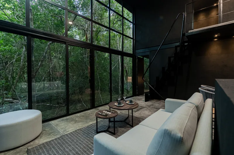 High-angle view of a modern living and kitchen area in a Tree60 2-bedroom Essence Jungle Villa, featuring large windows overlooking lush jungle, a light-colored sofa, dark kitchen island with stools, and concrete-style flooring.