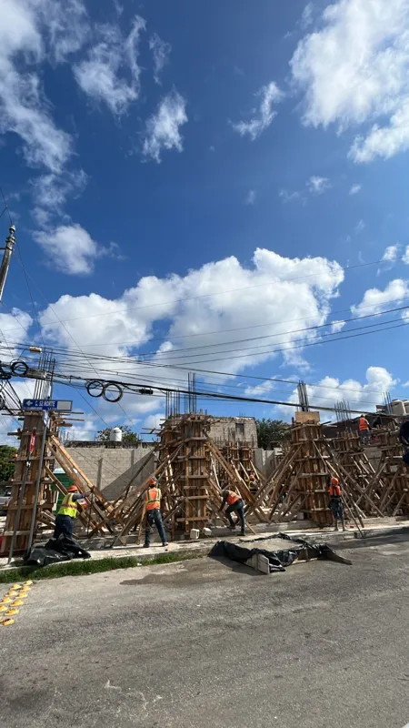 A wide shot of the Spirit 1990 construction site under a bright blue sky with white clouds. Several construction workers in orange safety vests and hard hats are visible, working on the building's foundation and ground floor. Extensive wooden form...