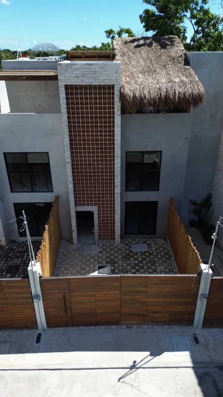 An aerial view of the front exterior of a multi-story house at Tamarindos development in Bacalar, featuring a large wooden gate, a paved courtyard, grey stucco walls, dark windows, a vertical decorative lattice panel, and a traditional thatched pa...