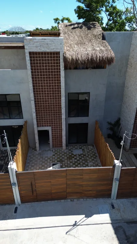 An aerial view of the front exterior of a multi-story house at Tamarindos development in Bacalar, featuring a large wooden gate, a paved courtyard, grey stucco walls, dark windows, a vertical decorative lattice panel, and a traditional thatched pa...