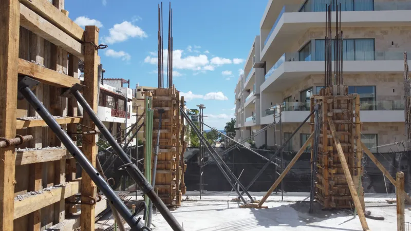 An aerial view of the Costera Mamitas construction site in Playa del Carmen, showing multiple workers in safety gear on a partially completed concrete slab, surrounded by wooden formwork for new columns and exposed rebar, with the vibrant blue Car...