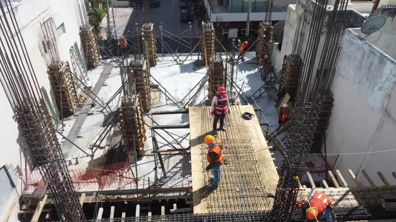 An aerial view of the Costera Mamitas construction site in Playa del Carmen, showing multiple workers in safety gear on a partially completed concrete slab, surrounded by wooden formwork for new columns and exposed rebar, with the vibrant blue Car...