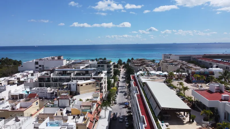 An aerial view of the Costera Mamitas construction site in Playa del Carmen, showing multiple workers in safety gear on a partially completed concrete slab, surrounded by wooden formwork for new columns and exposed rebar, with the vibrant blue Car...