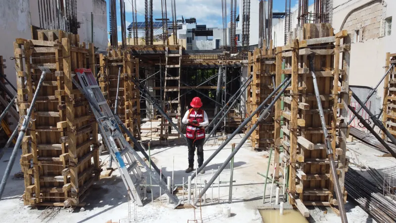 An aerial view of the Costera Mamitas construction site in Playa del Carmen, showing multiple workers in safety gear on a partially completed concrete slab, surrounded by wooden formwork for new columns and exposed rebar, with the vibrant blue Car...