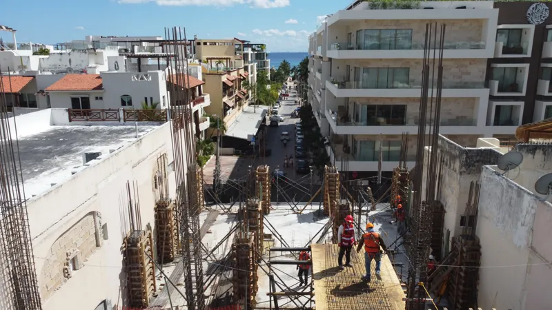 An aerial view of the Costera Mamitas construction site in Playa del Carmen, showing multiple workers in safety gear on a partially completed concrete slab, surrounded by wooden formwork for new columns and exposed rebar, with the vibrant blue Car...