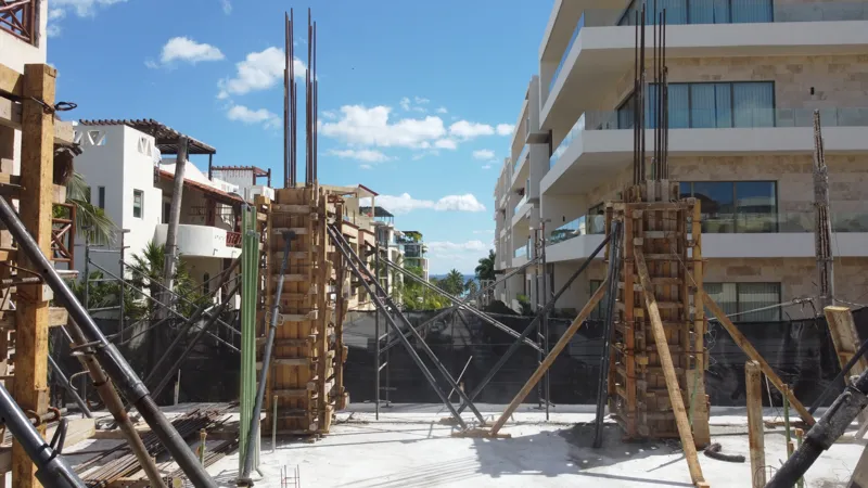 An aerial view of the Costera Mamitas construction site in Playa del Carmen, showing multiple workers in safety gear on a partially completed concrete slab, surrounded by wooden formwork for new columns and exposed rebar, with the vibrant blue Car...